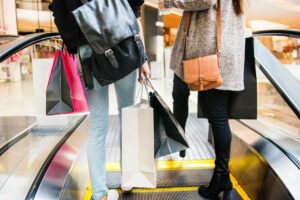 Duas mulheres em uma viagem de compras, subindo uma escada rolante, vestindo moda de rua e carregando bolsas.